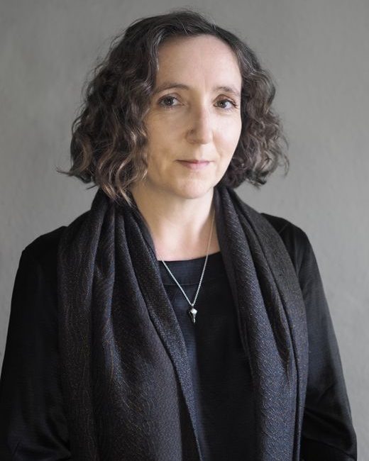 portrait of a white woman with mid brown curly hair to shoulder length. She is wearing a black dress and scarf